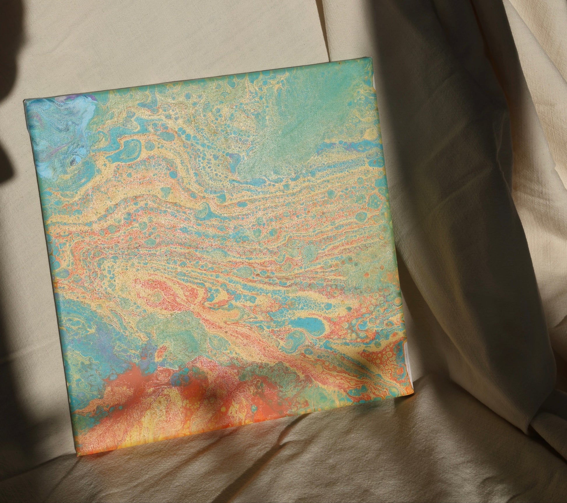 Abstract painting in 30x30 cm format with fluid patterns in blue, green, orange and beige tones, displayed against soft beige fabric by Lily Glow Decor