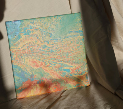 Abstract painting in 30x30 cm format with fluid patterns in blue, green, orange and beige tones, displayed against soft beige fabric by Lily Glow Decor