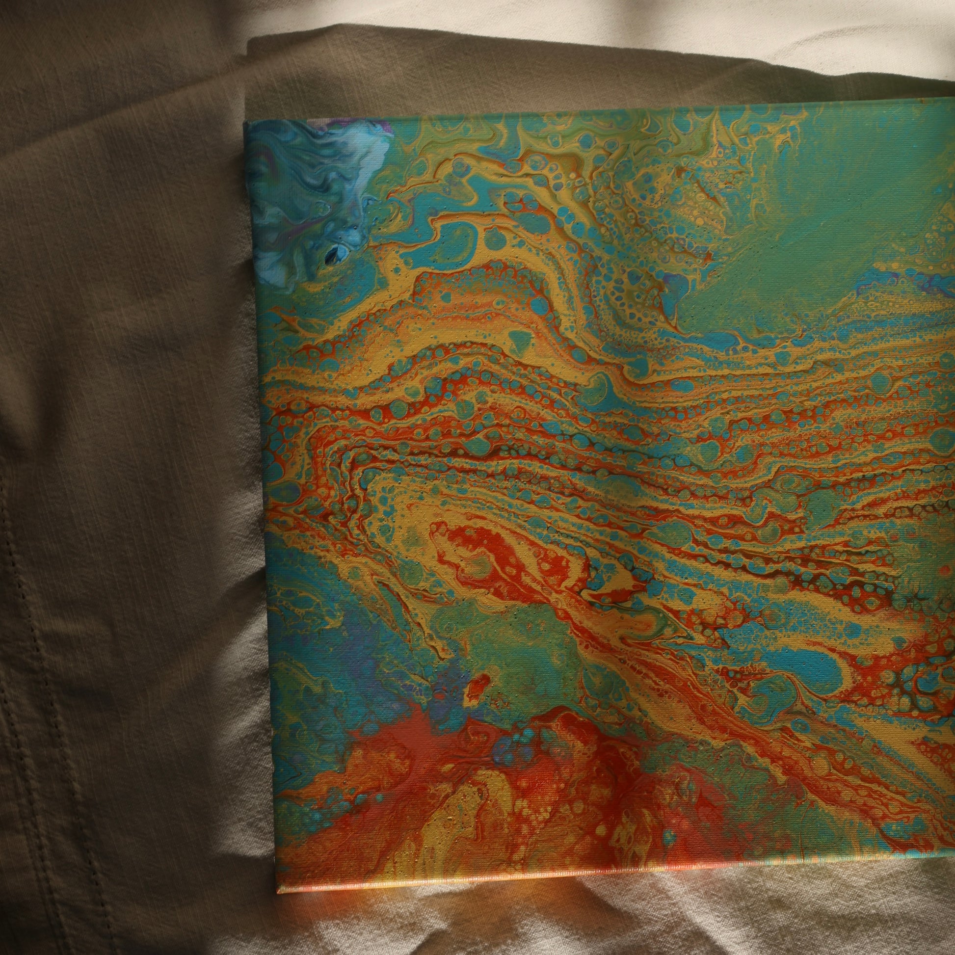 Abstract painting in 30x30 cm format with fluid patterns in blue, green, orange and beige tones, displayed against soft beige fabric by Lily Glow Decor